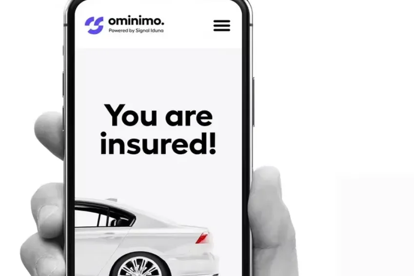 A hand holds a smartphone displaying “You are insured!” above a white car on an insurance webpage.