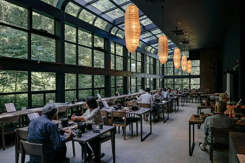 Diners sit at wooden tables in a restaurant with glowing hanging lanterns and tall windows overlooking leafy trees.